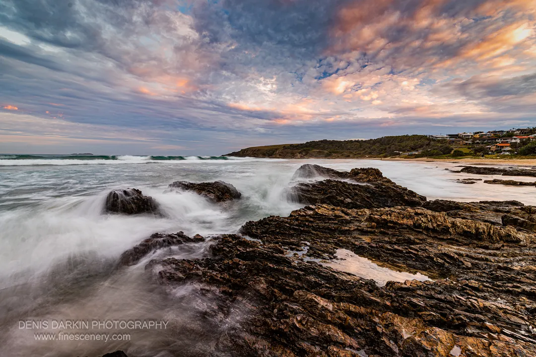 Long-exposure waves wash over a dark basalt reef near Coffs Harbor, tidal pools reflecting the pink dusk sky, a sandy cove in the distance.