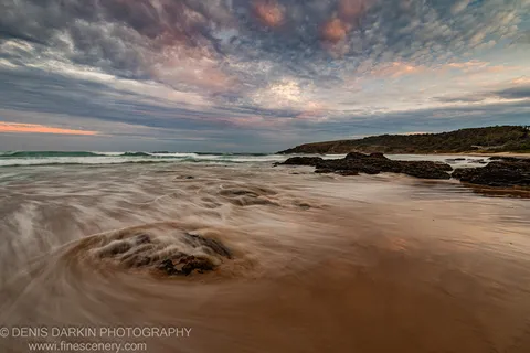 Smooth long-exposure surf over dark reef and wet sand on the New South Wales coast, swirling water in the foreground.