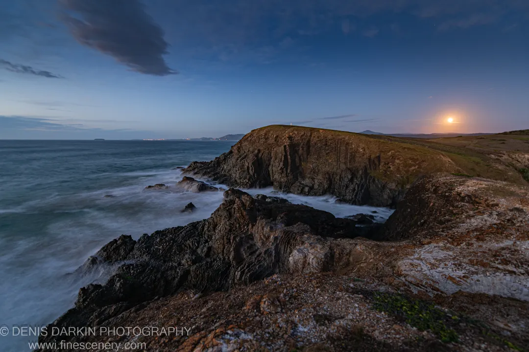 Rocky headland at Coffs Harbor at predawn, a full moon above the horizon and long-exposure waves blurred to mist below.