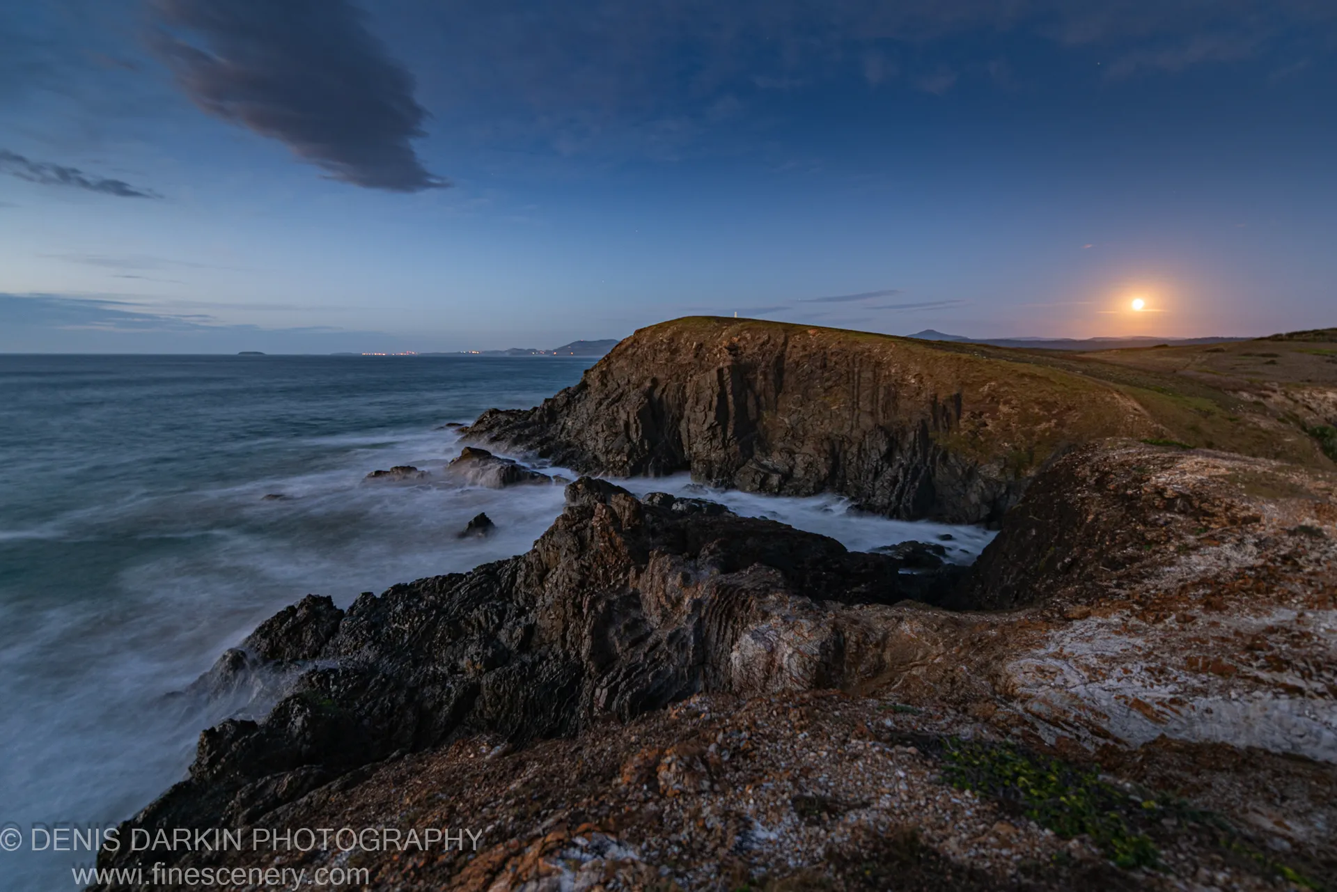 Rocky headland at Coffs Harbor at predawn, a full moon above the horizon and long-exposure waves blurred to mist below.