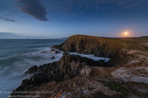 Rocky headland at Coffs Harbor at predawn, a full moon above the horizon and long-exposure waves blurred to mist below.