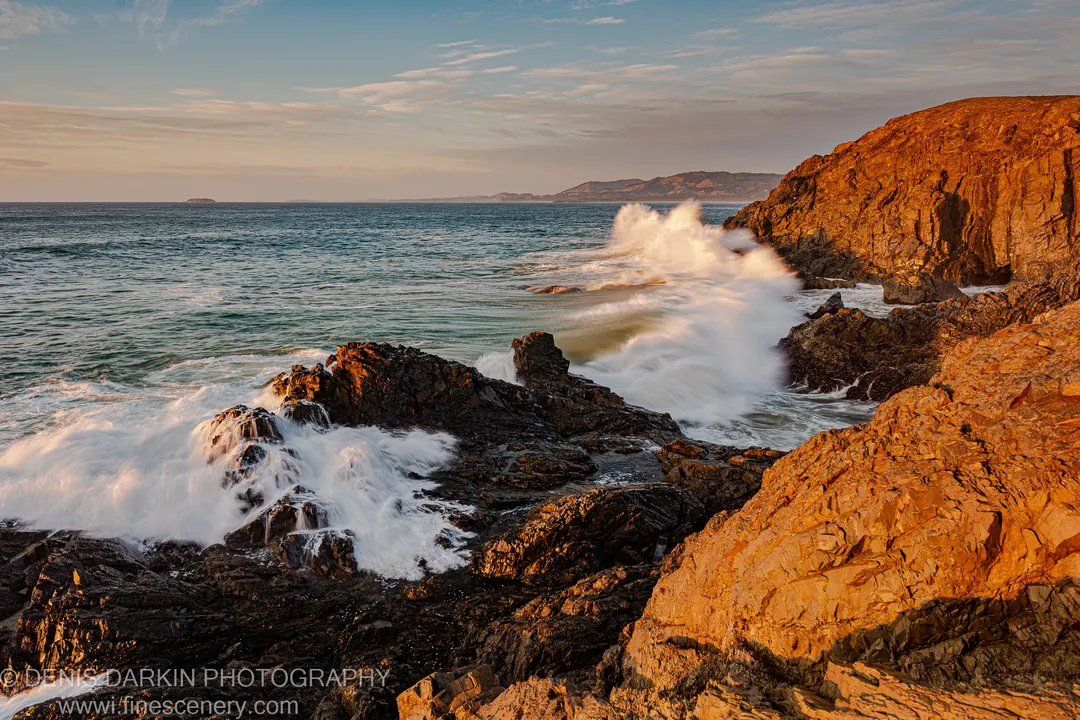 White surf crashes against dark basalt formations below sandstone cliffs at dawn on the Coffs Harbor coast, the cliff face in warm early light.