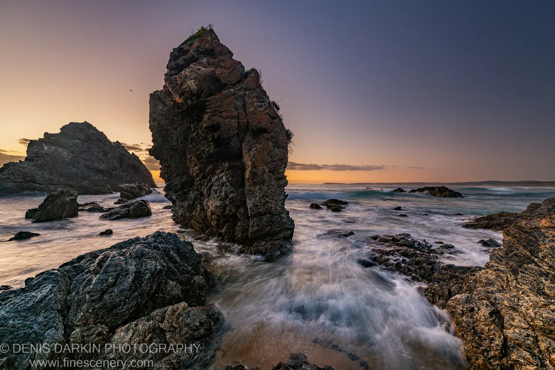 A tall sea stack on the South Coast at sunrise, with long-exposure waves reduced to white ribbons around dark foreground rocks.