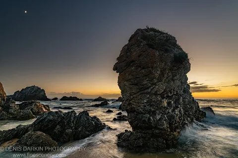 A stratified sea stack on the South Coast before dawn, with a crescent moon above the horizon and long-exposure surf.