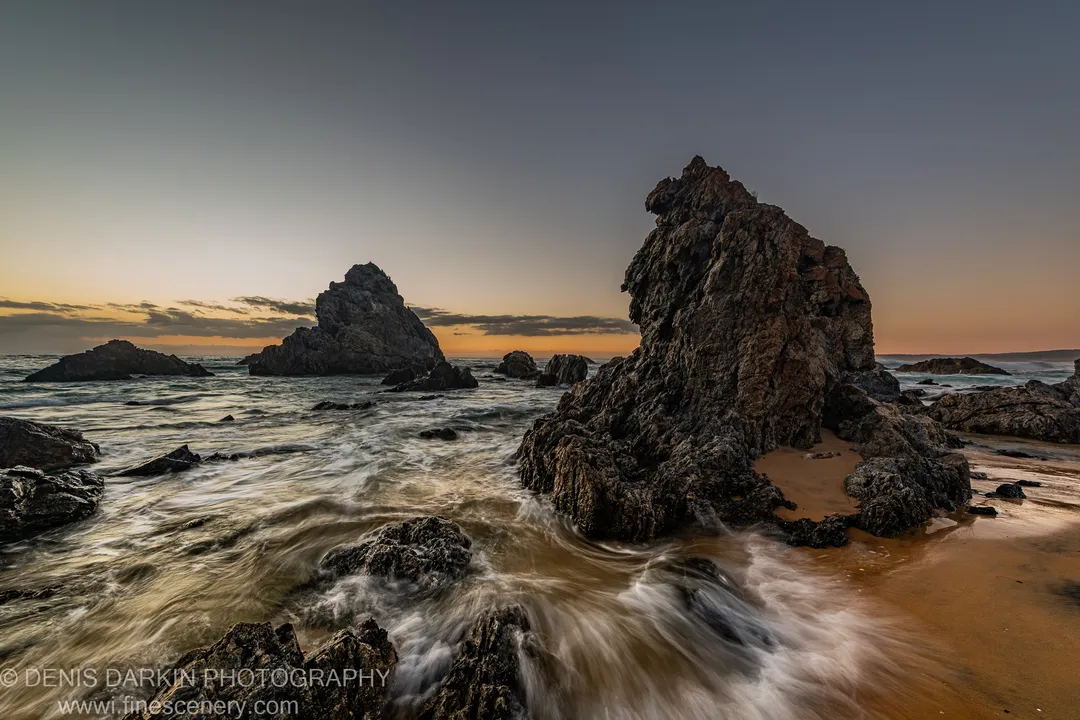 Jagged sea stacks at dawn on the South Coast, with a long-exposure tidal wash across the wet sand in the foreground.