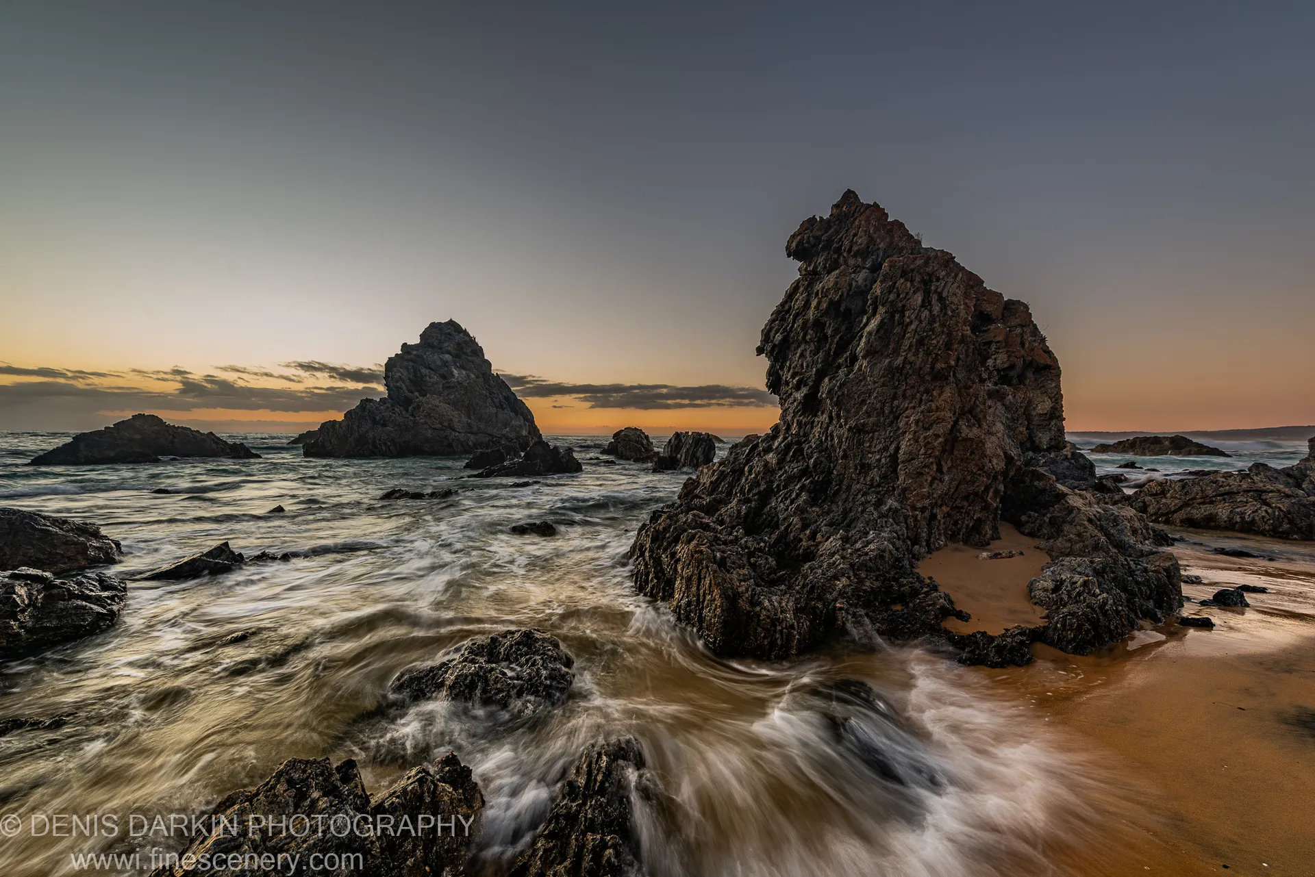 Jagged sea stacks at dawn on the South Coast, with a long-exposure tidal wash across the wet sand in the foreground.