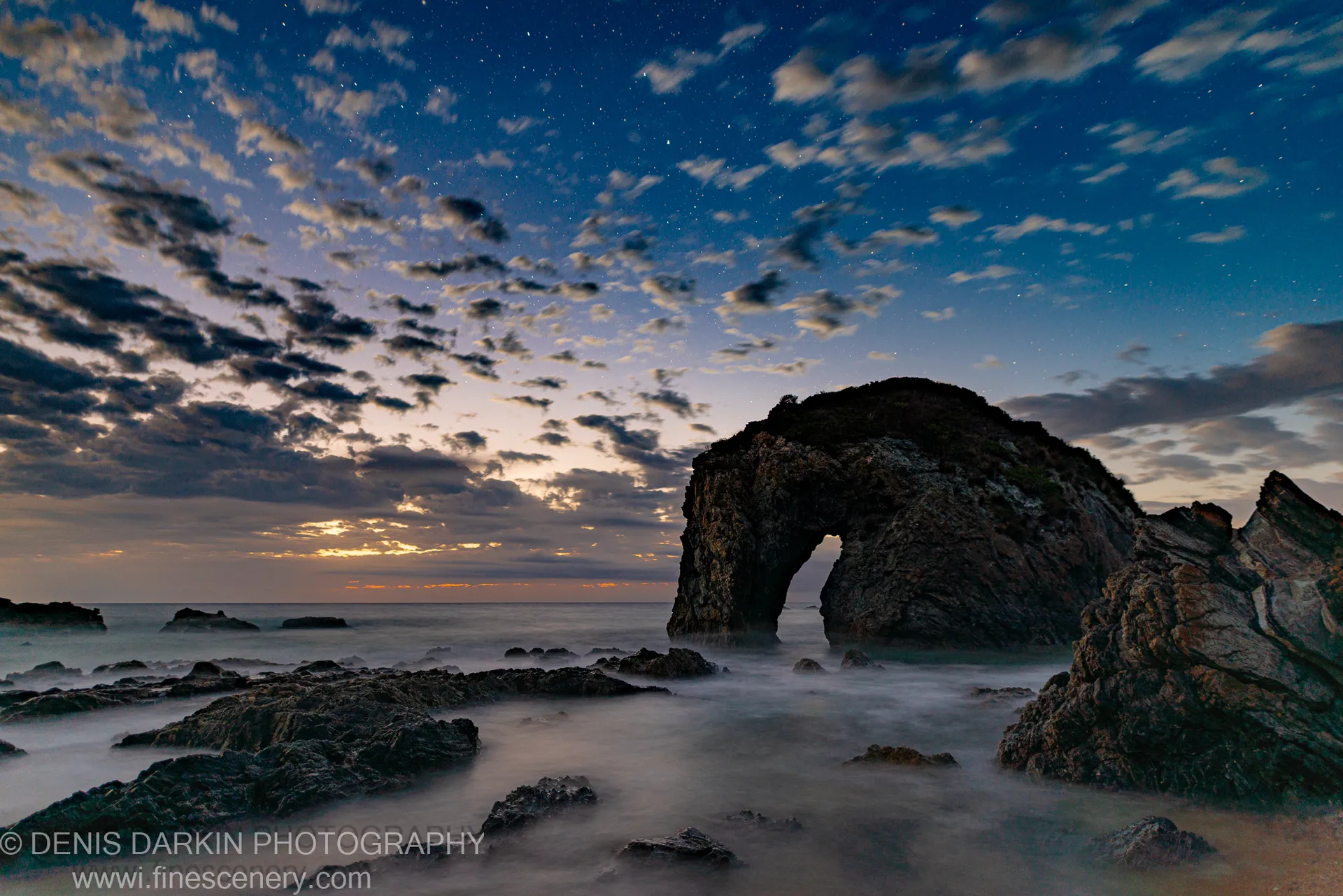 A rock arch on the Bermagui coast stands against a star-filled predawn sky, long-exposure waves softening around the rugged foreground rocks.