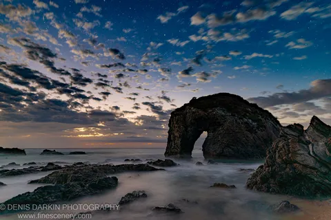 A rock arch on the Bermagui coast stands against a star-filled predawn sky, long-exposure waves softening around the rugged foreground rocks.