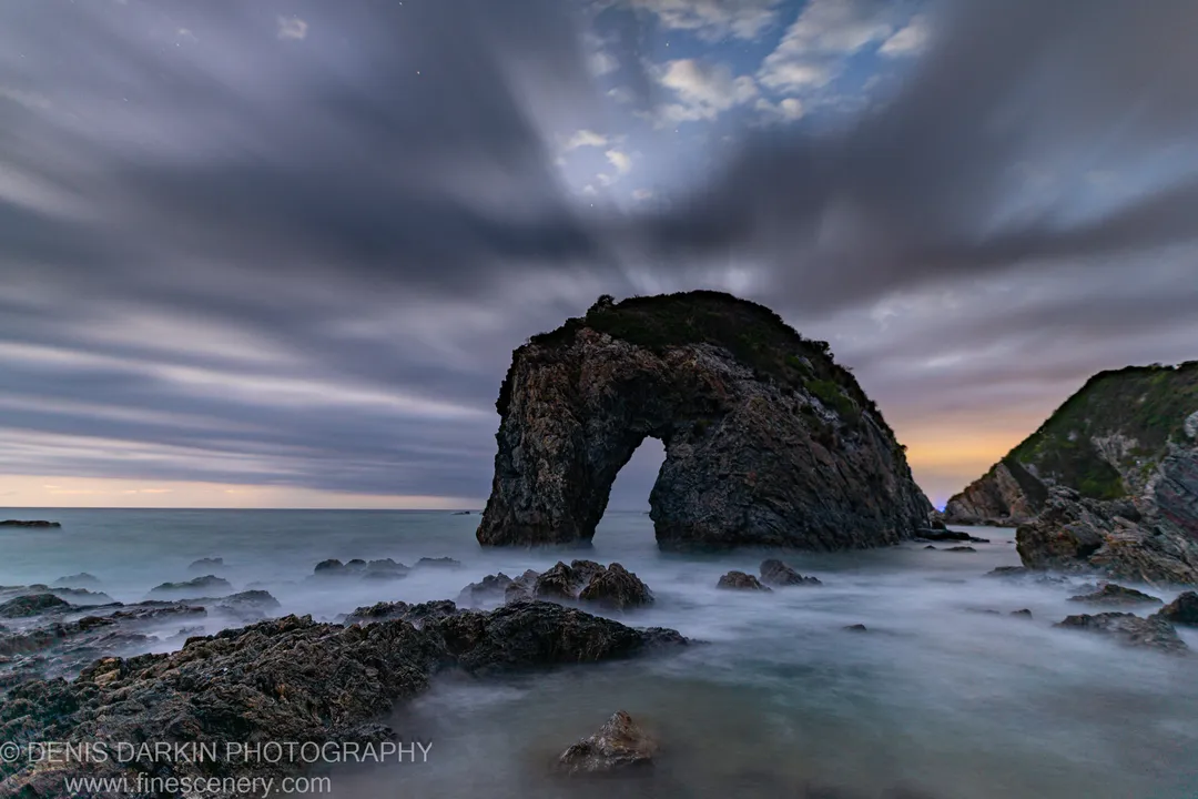 A natural rock arch before dawn on the South Coast, with long-exposure surf and streaked clouds under a deep blue sky.