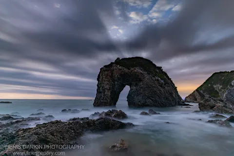 A natural rock arch before dawn on the South Coast, with long-exposure surf and streaked clouds under a deep blue sky.