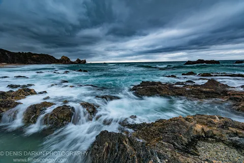 Storm clouds over dark rocks and turbulent surf on the South Coast at dawn, with sea stacks in the mid-ground.