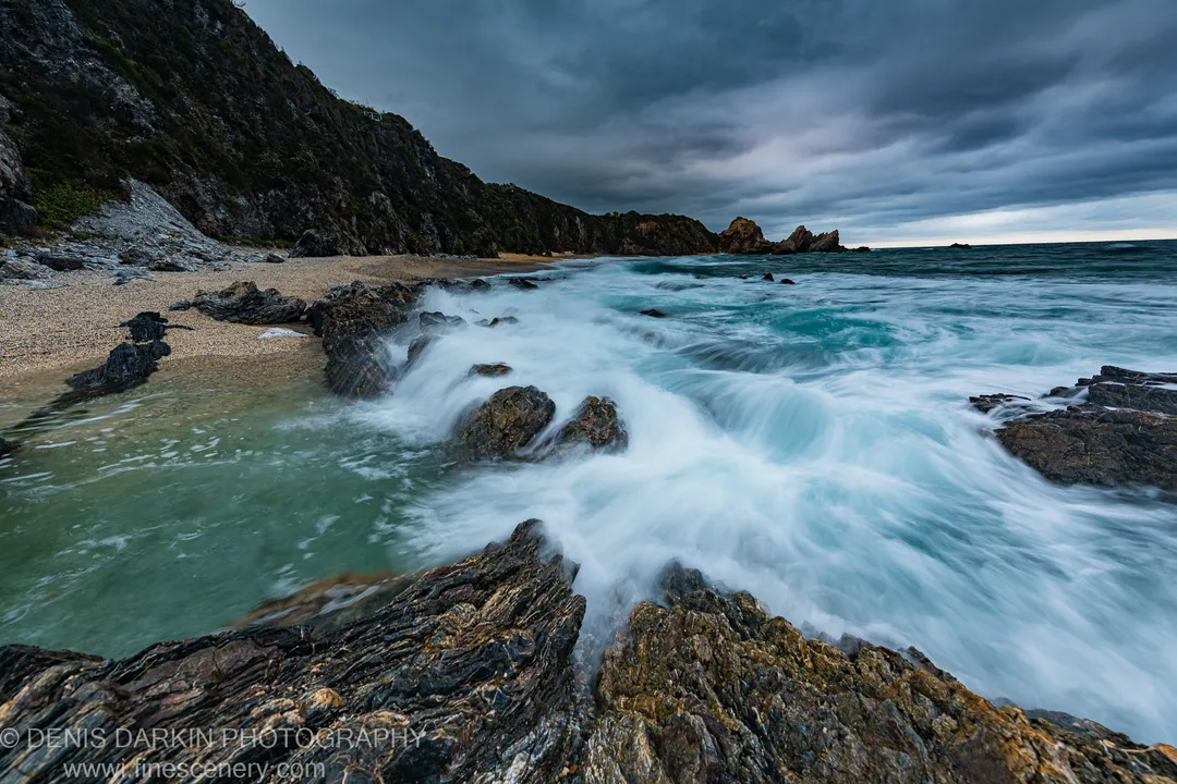Dark rocks and blurred wave motion lead toward a sandy cove and headland under heavy cloud, South Coast, New South Wales.