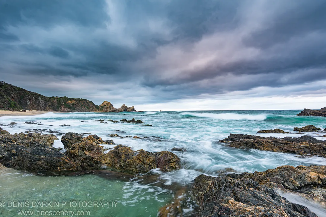Algae-covered rocks with turquoise surf and sea stacks behind, under heavy cloud on the South Coast at dawn.