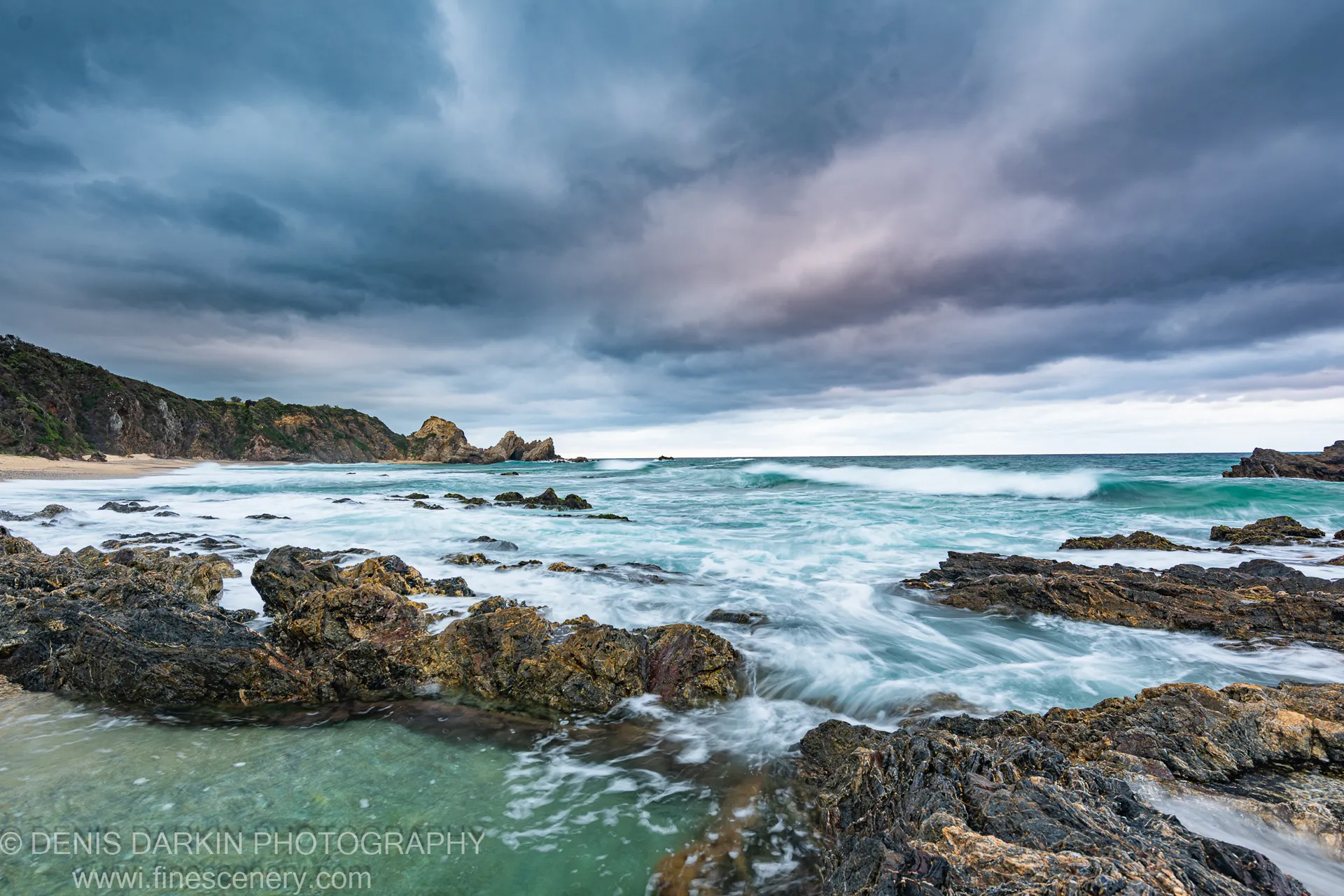 Algae-covered rocks with turquoise surf and sea stacks behind, under heavy cloud on the South Coast at dawn.
