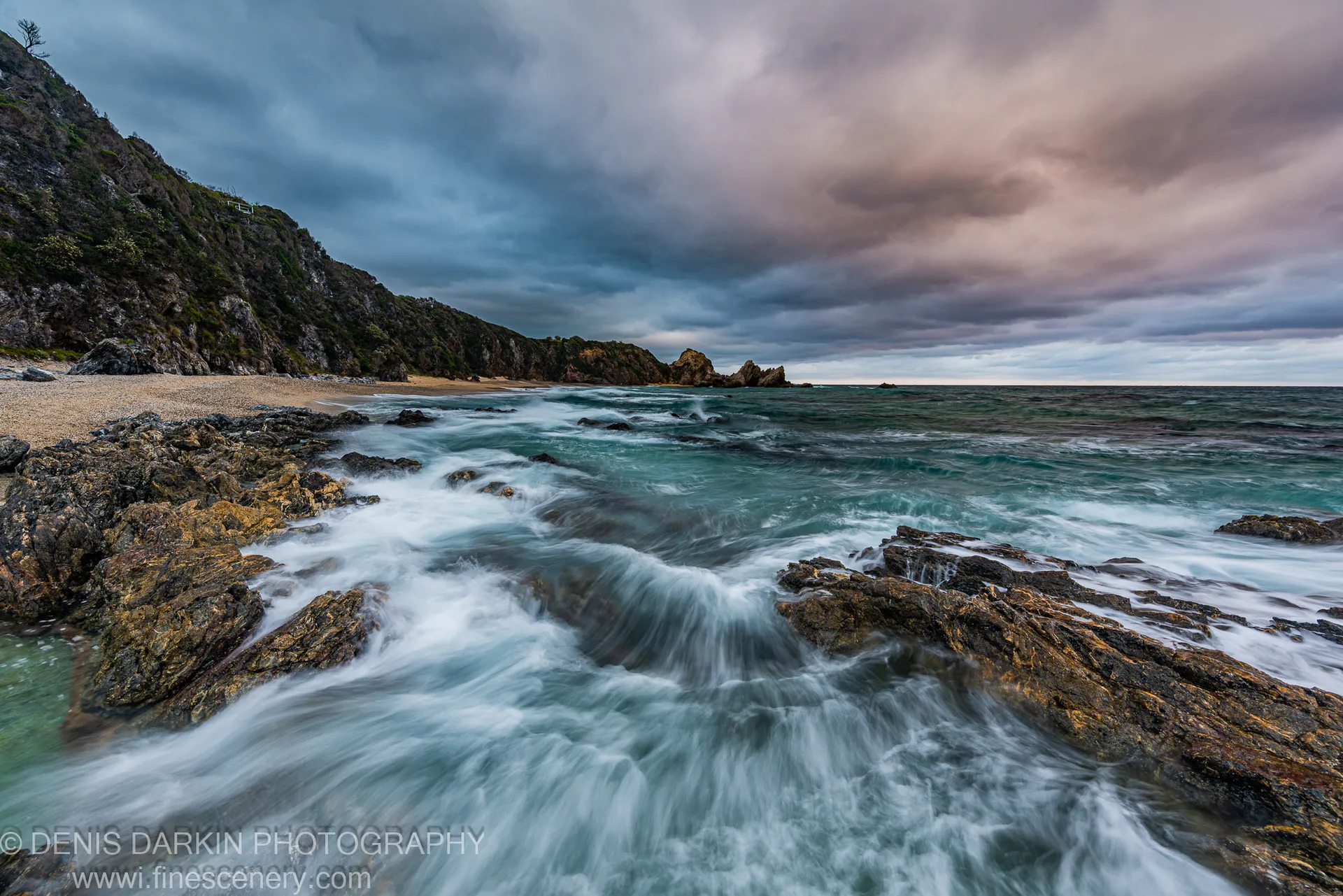 Long-exposure surf flows over lichen-covered rocks toward a sheltered cove on the South Coast of New South Wales at dawn.