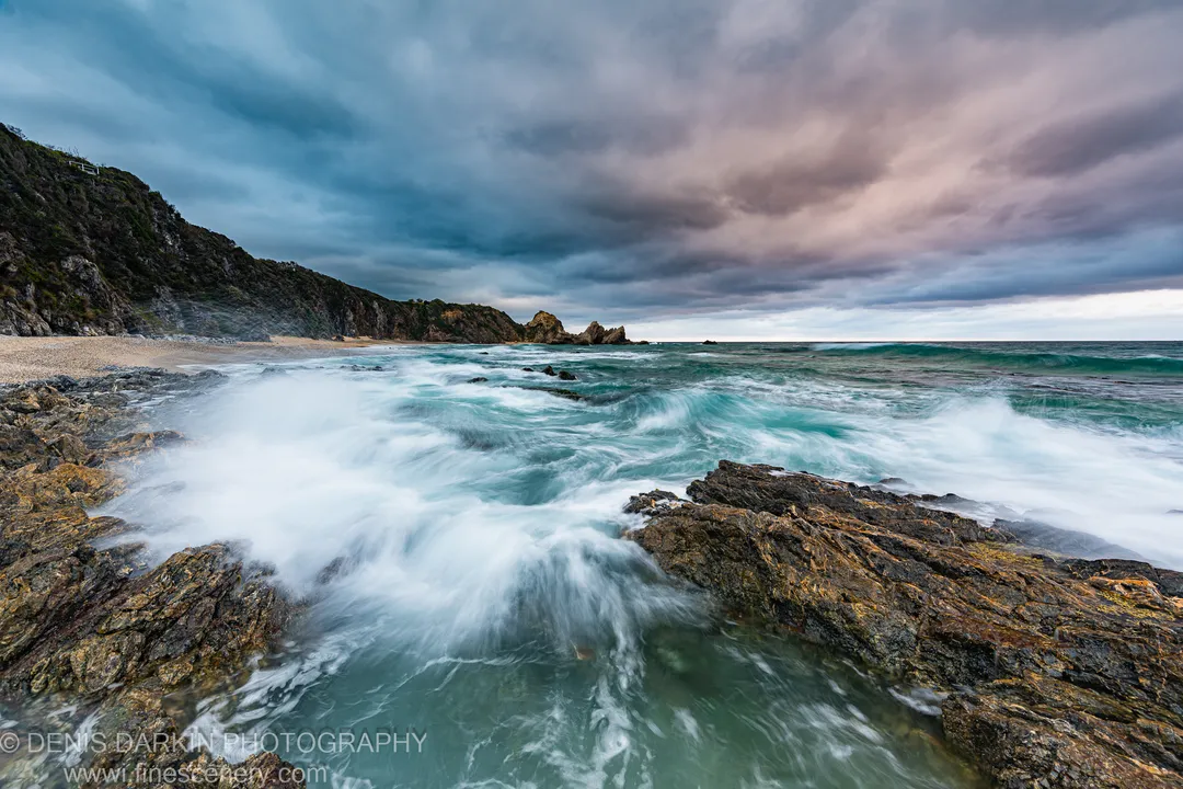 Long-exposure waves crash over lichen-stained rocks below a steep coastal cliff on the South Coast of New South Wales.