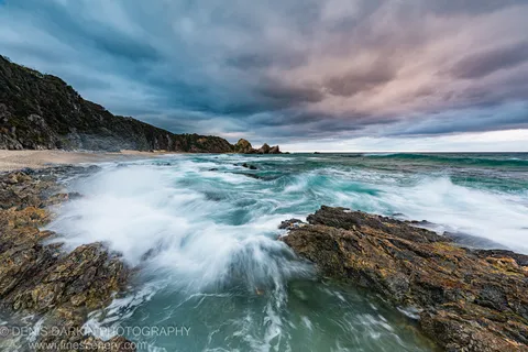 Long-exposure waves crash over lichen-stained rocks below a steep coastal cliff on the South Coast of New South Wales.