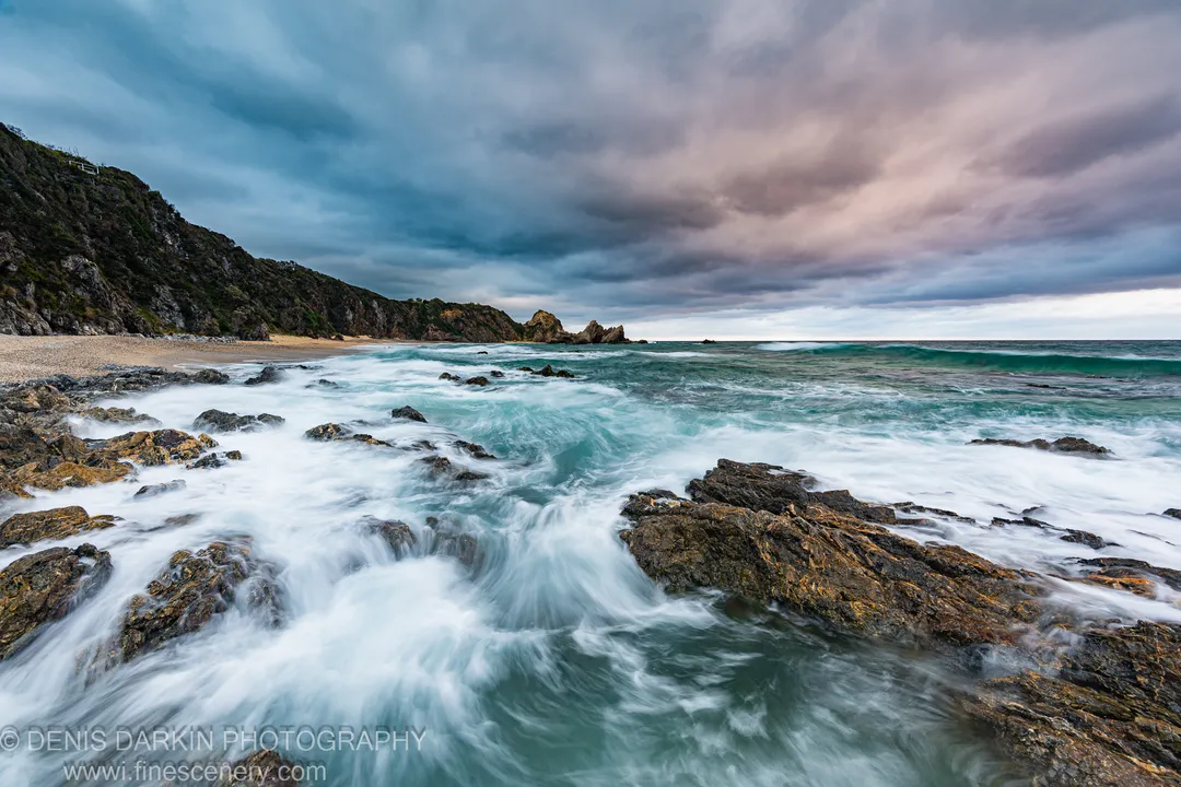 Long-exposure surf washes over rugged rock formations on the South Coast of New South Wales at dawn, with layered storm clouds above.