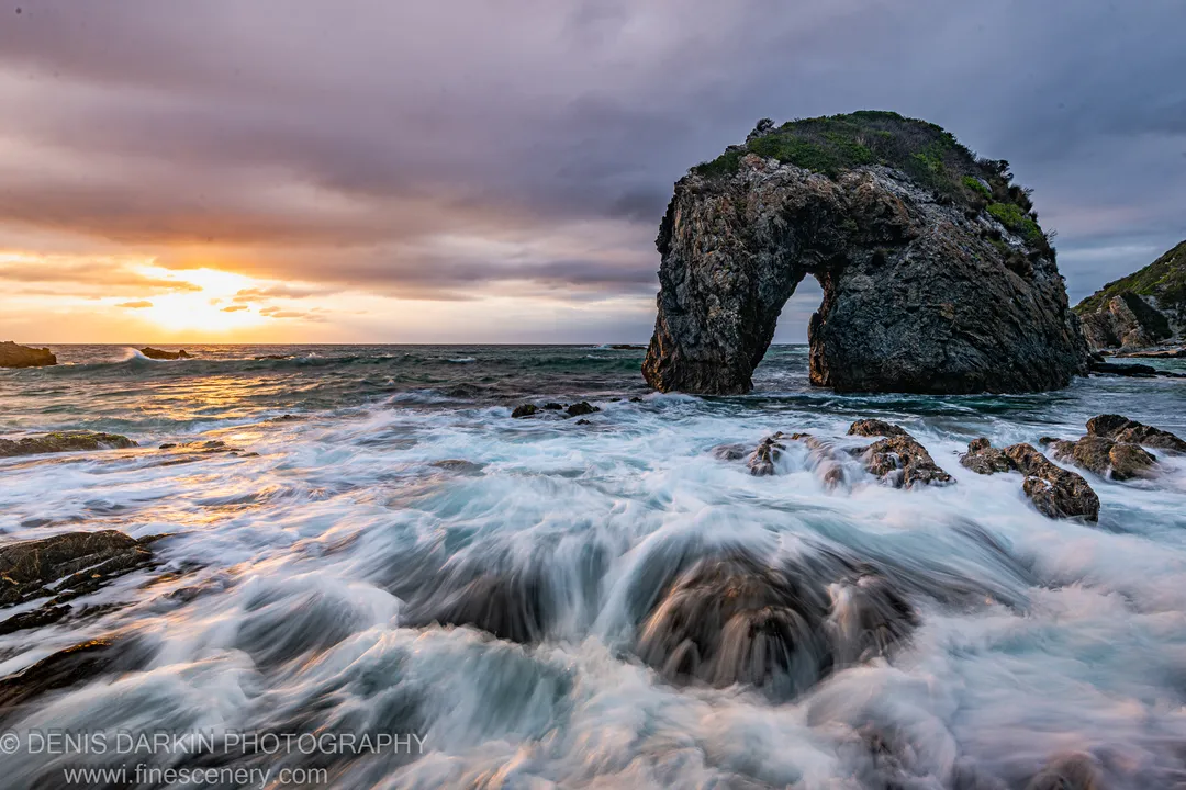 Long-exposure surf moves around dark foreground rocks below a natural sea arch silhouetted against storm clouds, New South Wales coast.