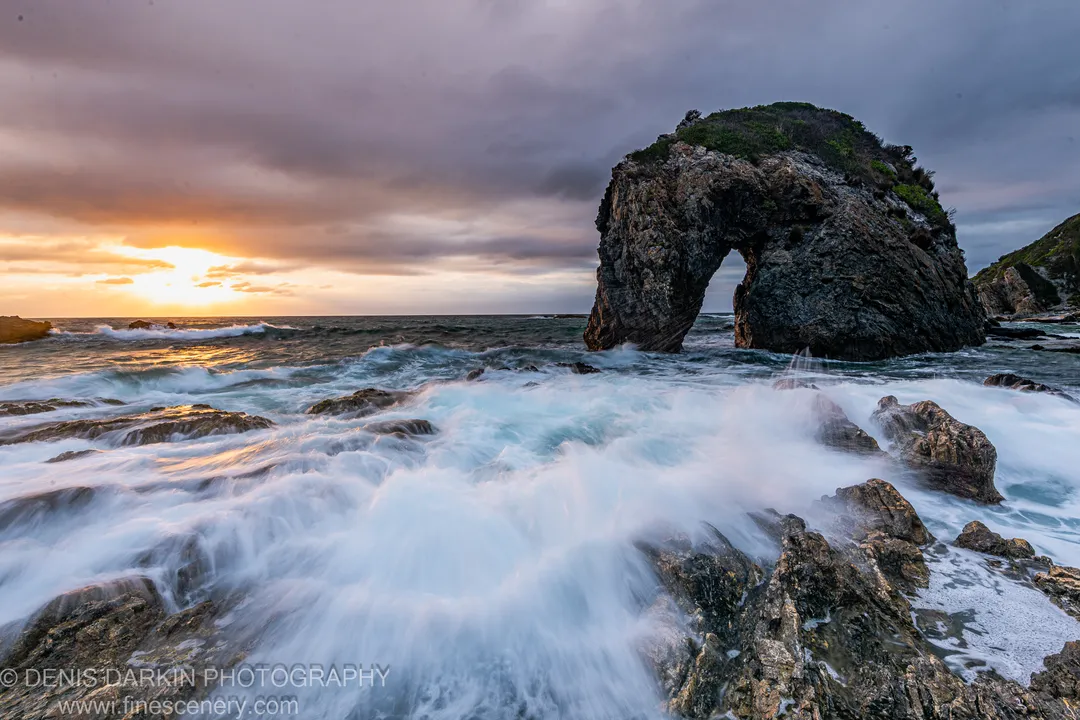 A sea stack with a natural arch stands above long-exposed surf breaking over jagged rocks on the South Coast of New South Wales.