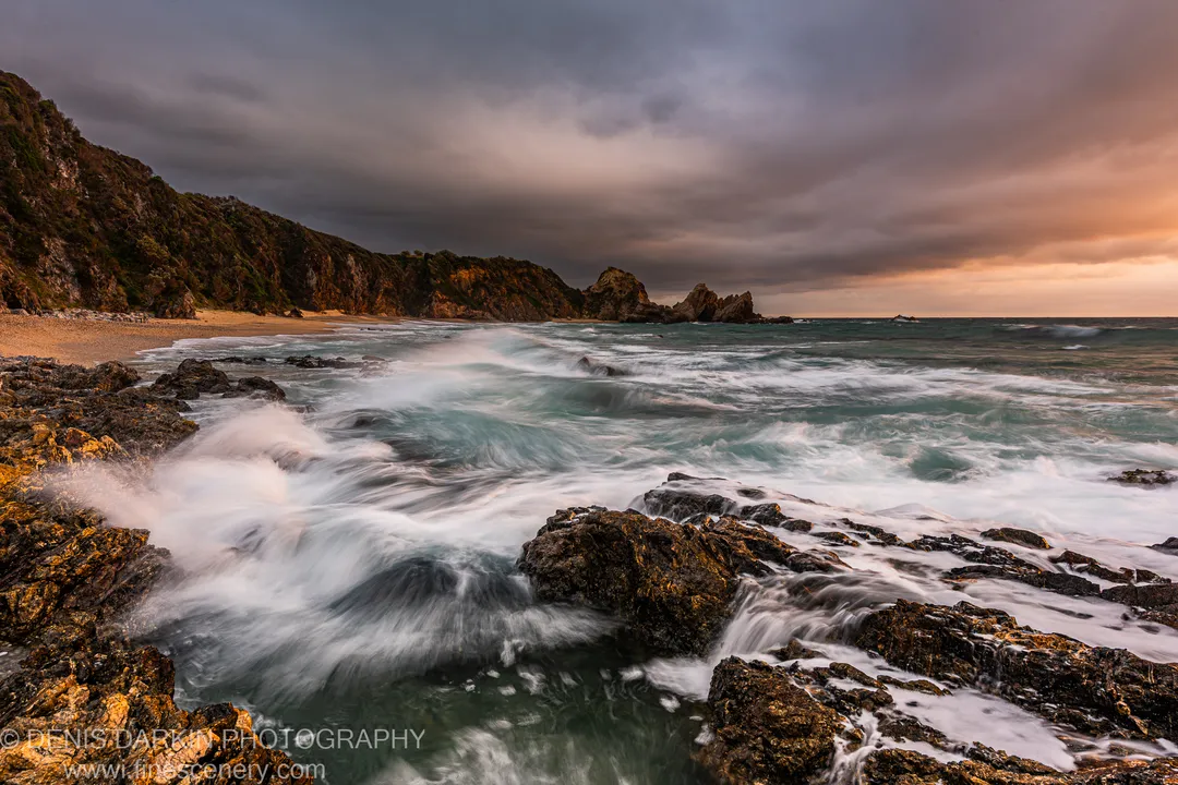 Dark algae-covered rocks in a sandy cove on the NSW South Coast, blurred waves catching a warm amber glow beneath storm clouds at dawn.