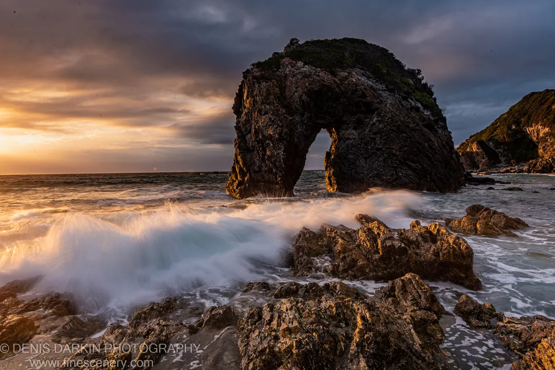 Warm dawn light catches the underside of a natural arch as long-exposure waves break around boulders on the New South Wales coast.