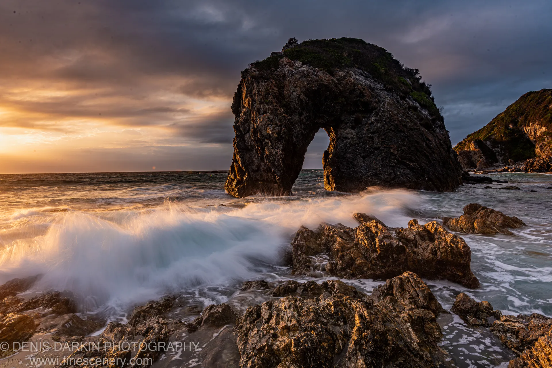 Warm dawn light catches the underside of a natural arch as long-exposure waves break around boulders on the New South Wales coast.