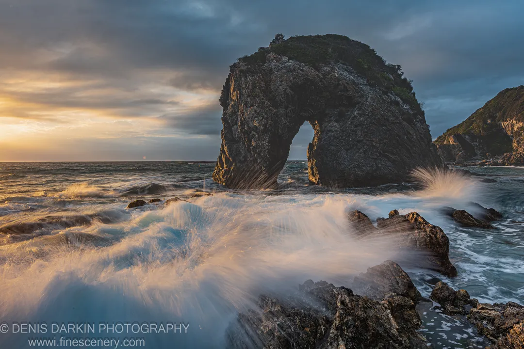 A massive sea stack with a natural arch rises above long-exposure surf and jagged foreground rocks on the South Coast, New South Wales.