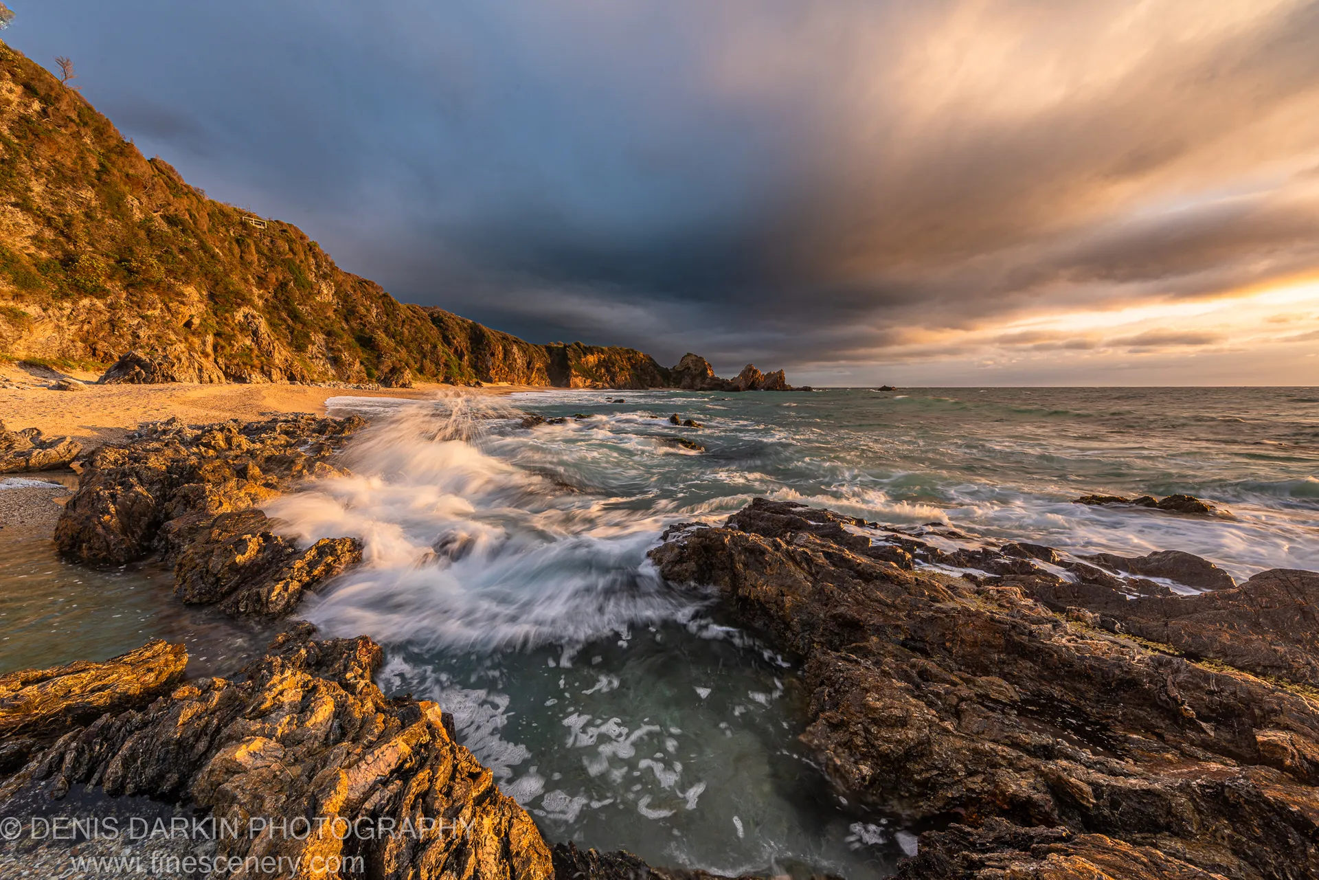 Basalt rocks and a sandy cove at dawn on the Bermagui coast, long-exposure waves blurred to mist beneath storm clouds turning orange at the horizon.