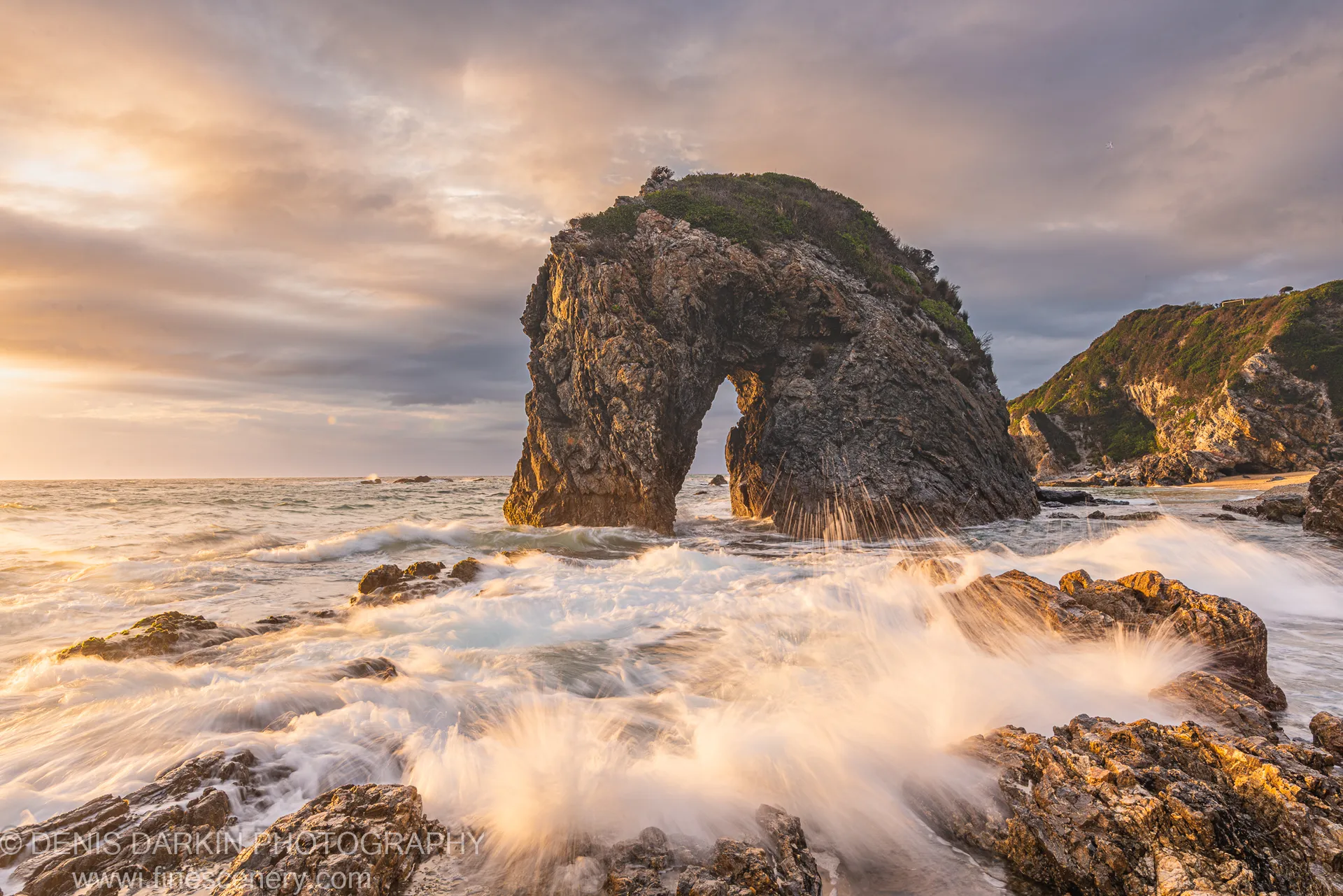 A rock arch on the Bermagui coast at dawn, surf crashing around dark foreground rocks below textured clouds with warm sunrise light at the horizon.