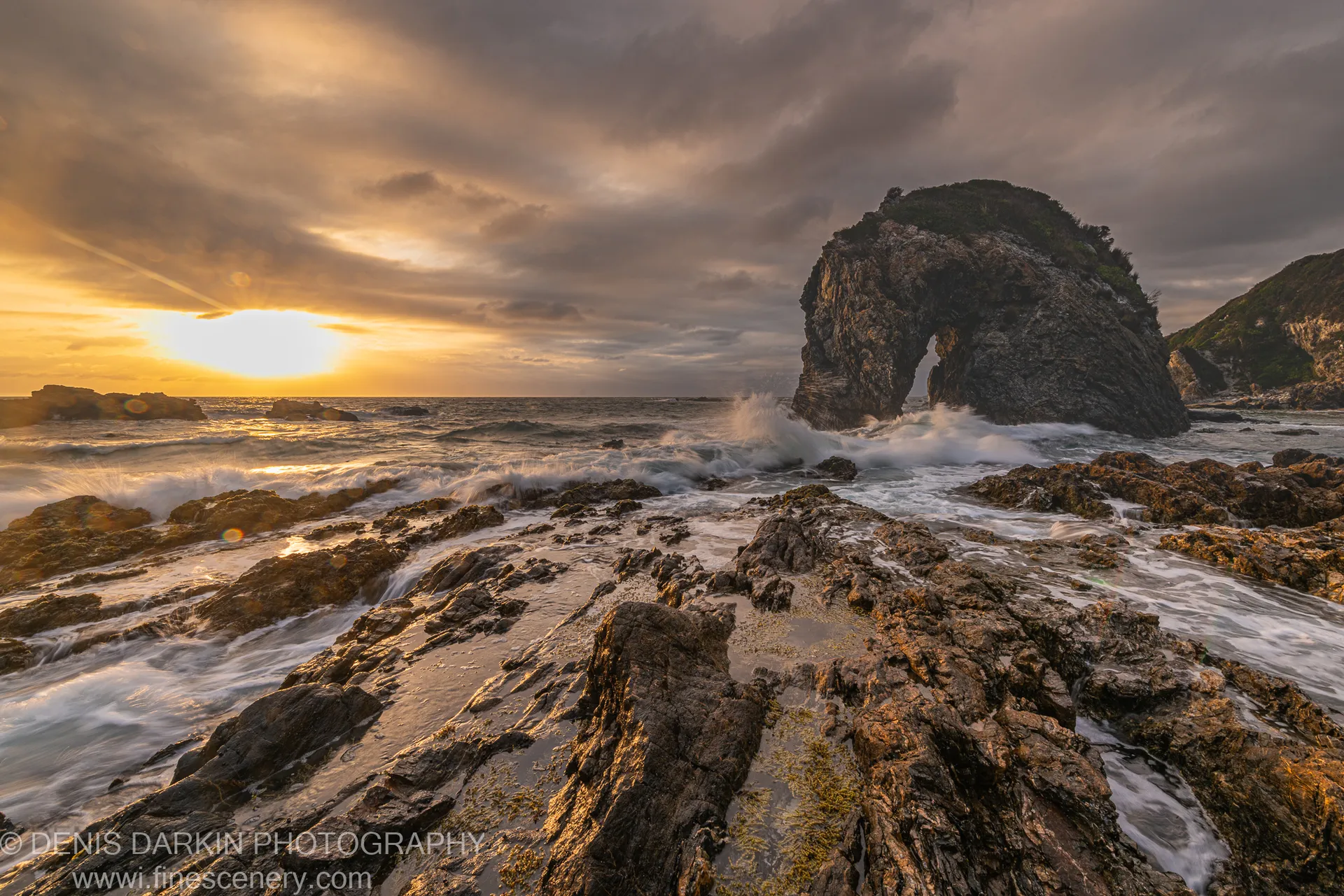 A rock arch on the Bermagui coast at dawn, spray from breaking waves lit by warm golden light from the left, heavy clouds overhead.