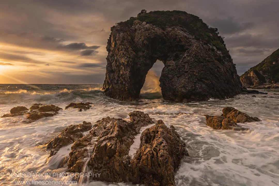 A sea stack with a natural arch on the Bermagui coast at dawn, storm-cloud light above surf swirling around wet foreground rocks.