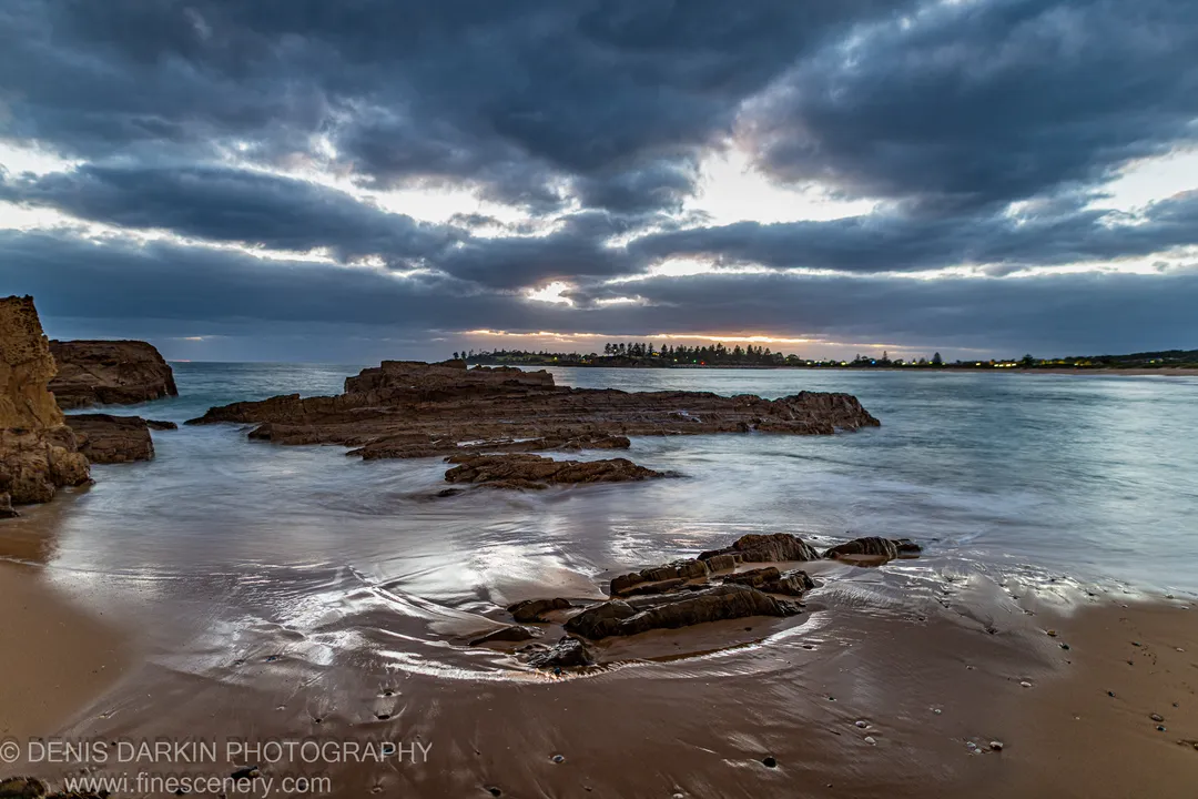 Storm clouds over a rocky reef on the Bermagui coast at dawn, long-exposure water between stratified formations, a sliver of light at the horizon.