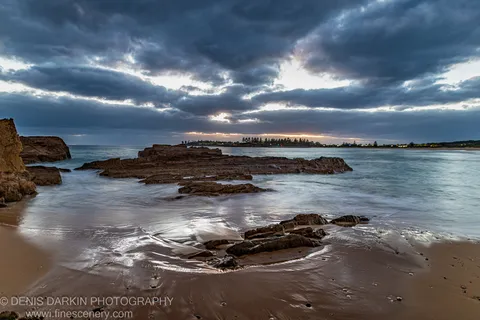 Storm clouds over a rocky reef on the Bermagui coast at dawn, long-exposure water between stratified formations, a sliver of light at the horizon.