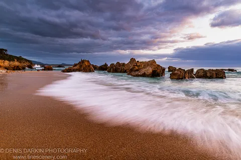 Jagged coastal rocks on the Bermagui coast at dawn, golden sand in the foreground, storm clouds above with warm light on the right horizon.