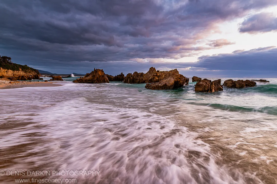 Foam-streaked waves sweep across wet sand toward a cluster of sea stacks beneath heavy storm clouds at Bermagui at dawn.