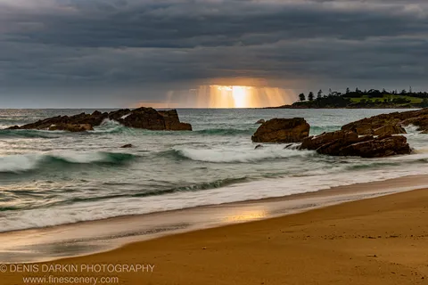 Crepuscular rays break through storm clouds above dark sandstone rocks and wave-washed sand on the NSW South Coast at dawn.