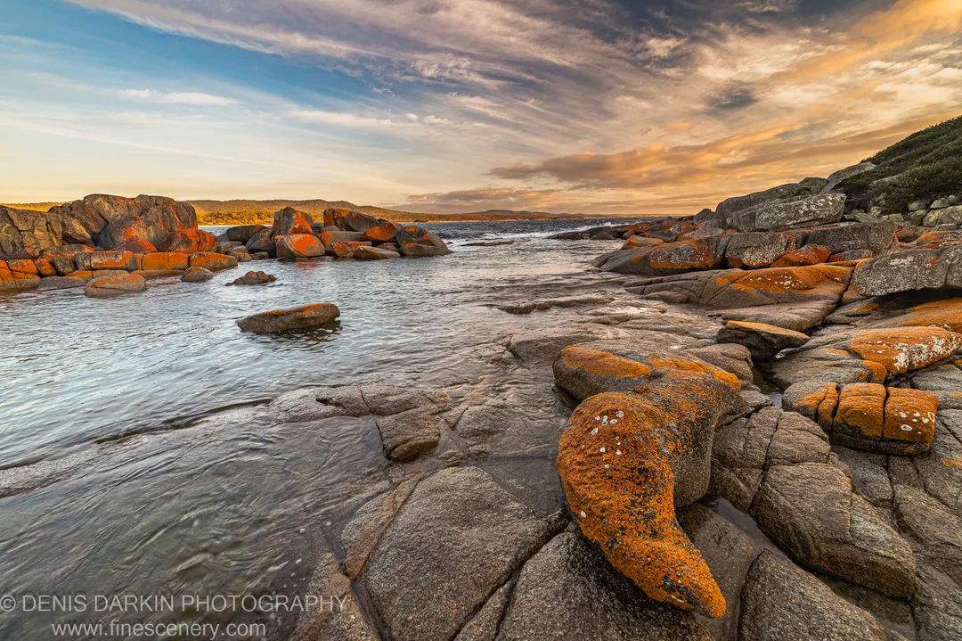 Golden light reflects off wet sand on the NSW South Coast as waves break around dark rocks, a headland silhouetted against storm clouds.