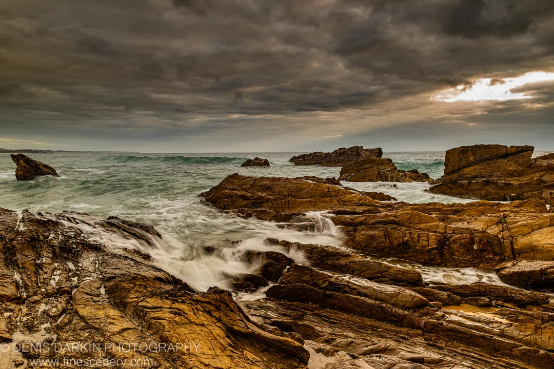 Layered sandstone shelves in warm amber tones meet green-grey waves under an overcast sky with a break of light, NSW South Coast.