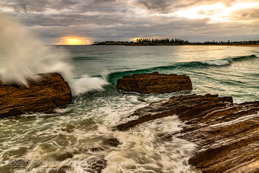 White sea spray rises against layered sandstone ledges on the NSW South Coast as a sunburst breaks through storm clouds over turquoise water.