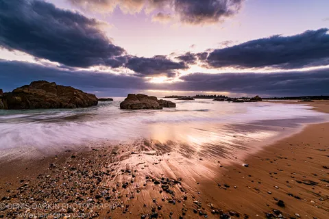 Long-exposure waves wash over wet sand beside coastal boulders at Bermagui as golden light breaks through storm clouds at dawn.
