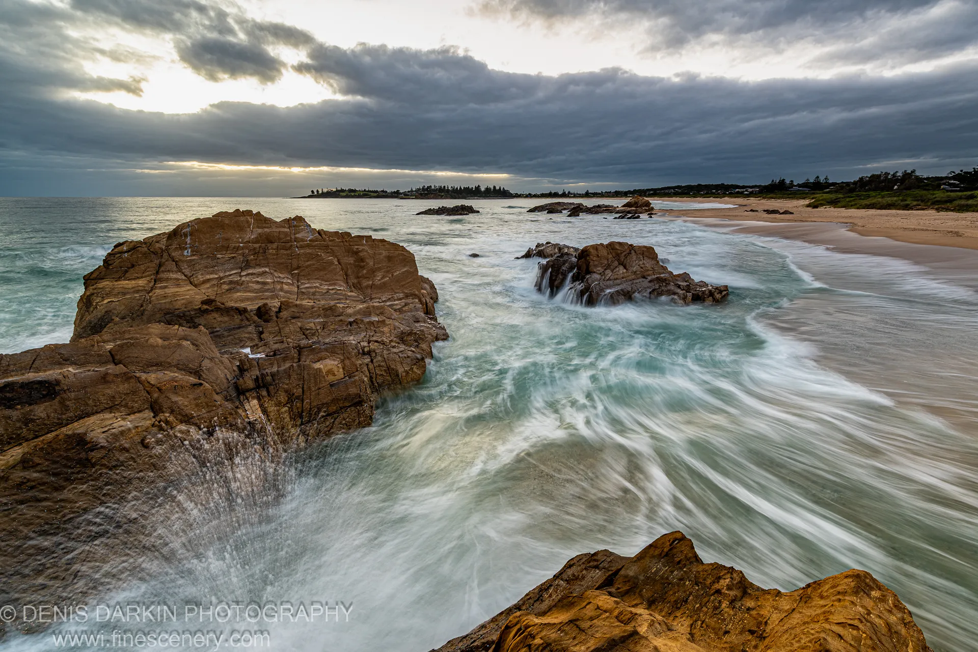 Layered sandstone formations in warm tones at Bermagui front a long-exposure sea below a stormy sky with a pale horizon break.