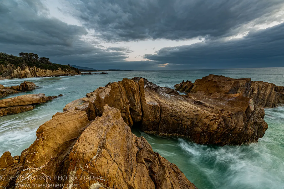 Broad sandstone ledges at Bermagui extend into a long-exposure sea beneath a low storm sky with a pale band of light near the horizon.