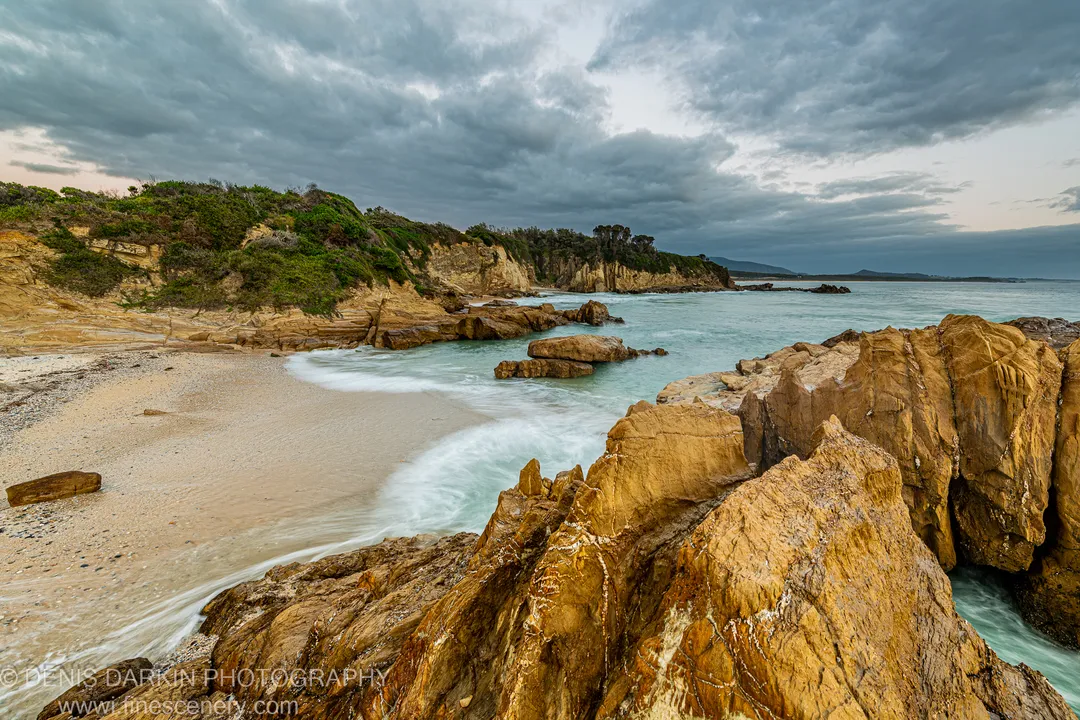 Honey-toned sandstone formations enclose a sheltered cove at Bermagui, with long-exposure waves softened into smooth water beneath a stormy sky.