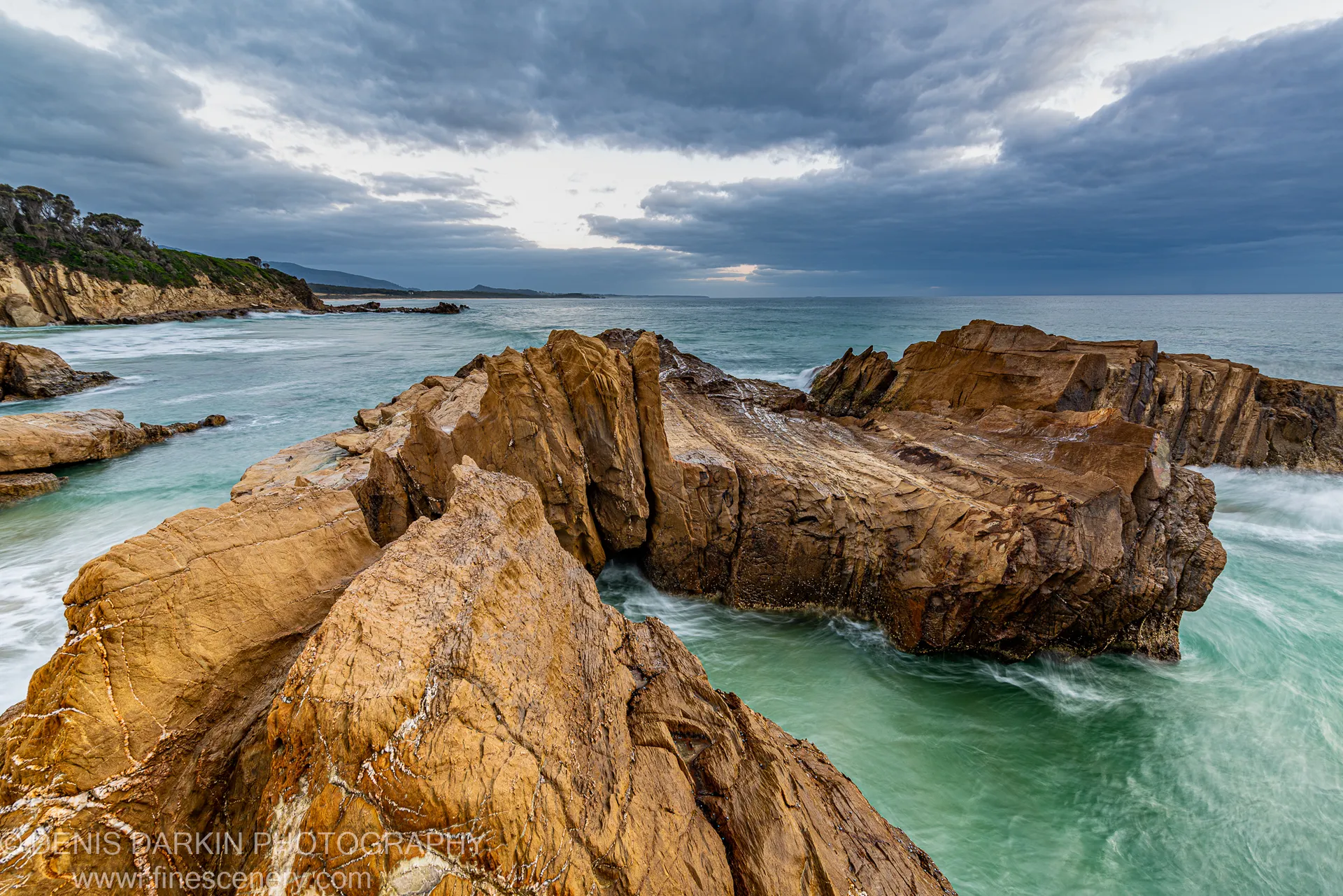 Honey-colored sandstone formations with vertical striations stand above a long-exposure sea at Bermagui, with a rugged headland visible in the distance.