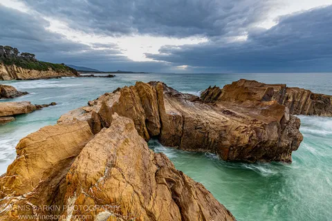 Honey-colored sandstone formations with vertical striations stand above a long-exposure sea at Bermagui, with a rugged headland visible in the distance.