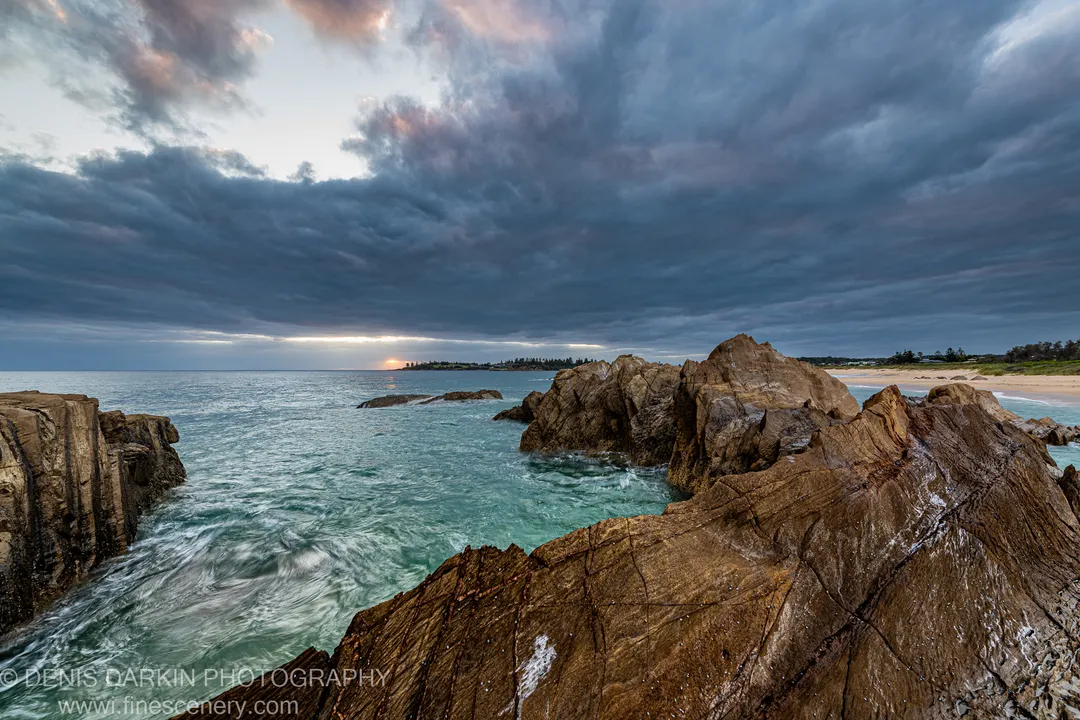 Wet, cracked rock with marine life in the foreground, turquoise water beyond, and a warm horizon glow under blue-grey clouds, NSW South Coast.