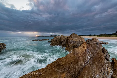 A massive sandstone formation stands above blurred surf at Bermagui, with storm clouds hanging low and a sliver of warm light at the horizon.