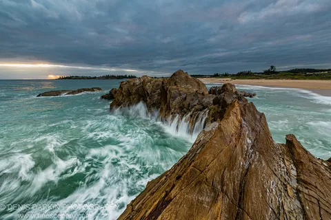 Striated rock formations jut into turquoise water at Bermagui, long-exposure waves blurred into soft ribbons below heavy clouds and a warm horizon break.