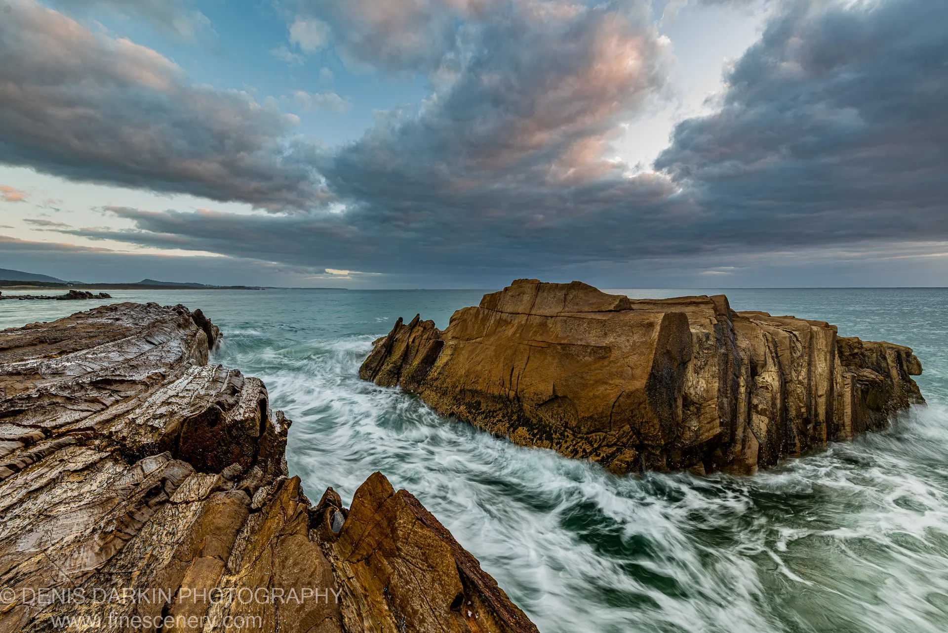 Layered rock outcrops at Bermagui protrude into deep water as long-exposure surf blurs around them below a soft pink and grey dawn sky.
